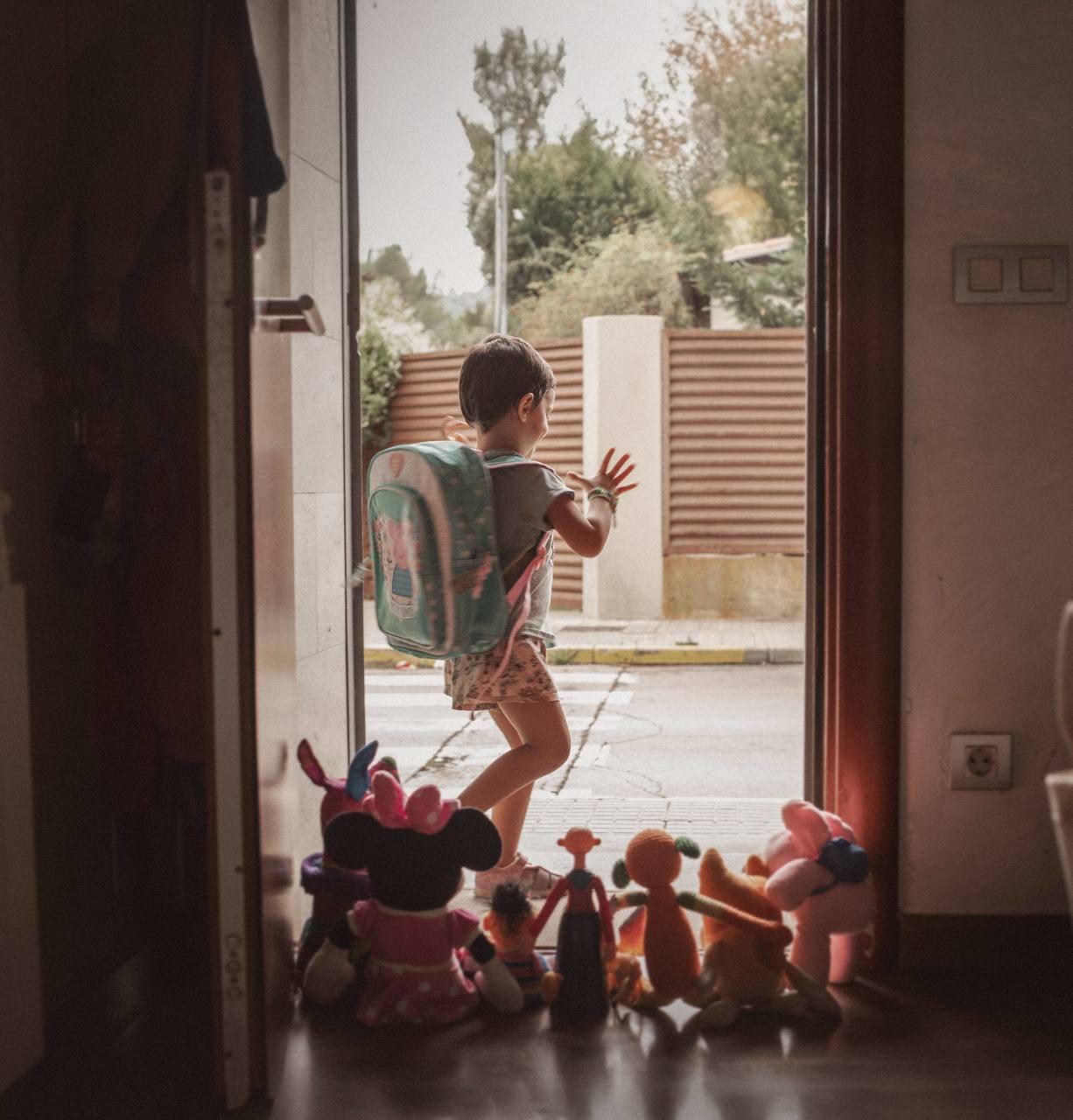 Una niña con mochila sale por la puerta de casa mientras sus peluches se quedan mirando desde dentro