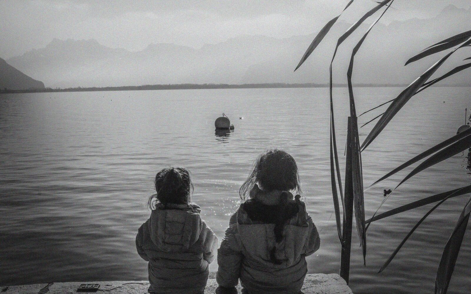 Dos niñas sentadas de espaldas mirando el agua de un lago en blanco y negro