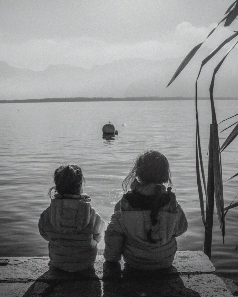 Dos niñas sentadas de espaldas mirando el agua de un lago en blanco y negro