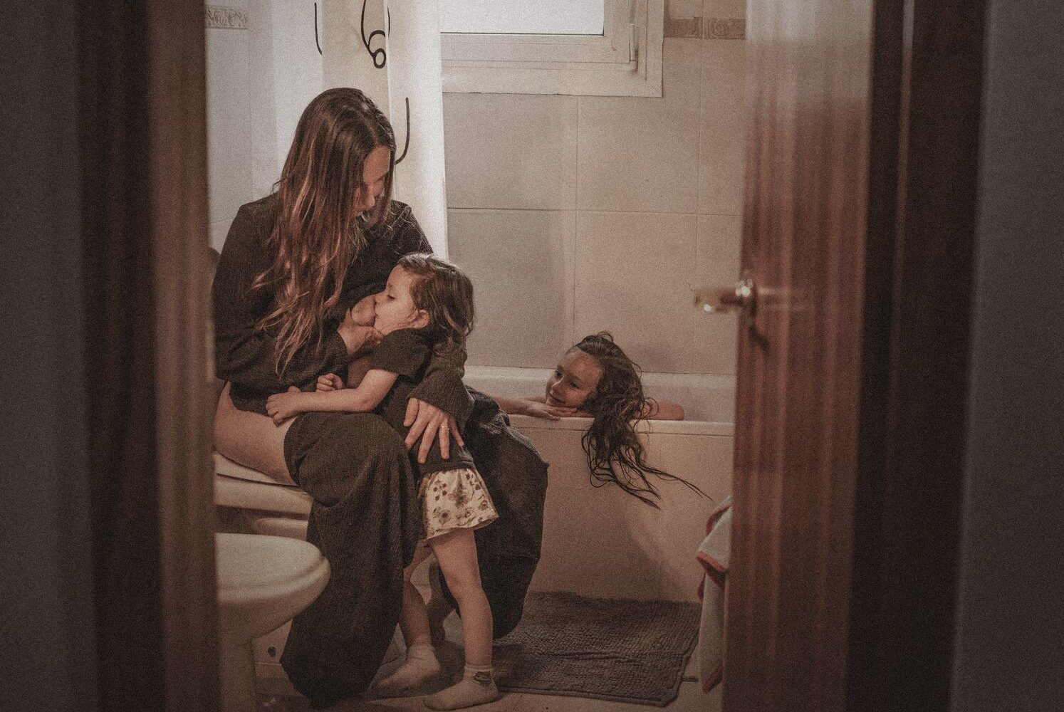 Una madre amamantando en el baño mientras su otra hija se baña, vista desde la puerta entreabierta