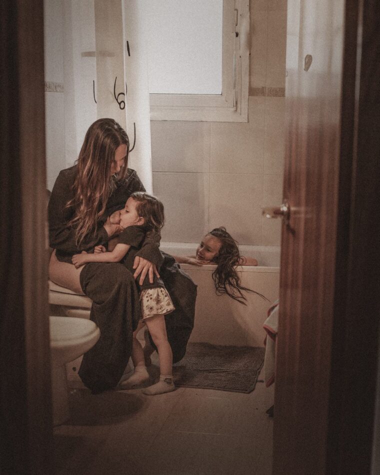 Una madre amamantando en el baño mientras su otra hija se baña, vista desde la puerta entreabierta