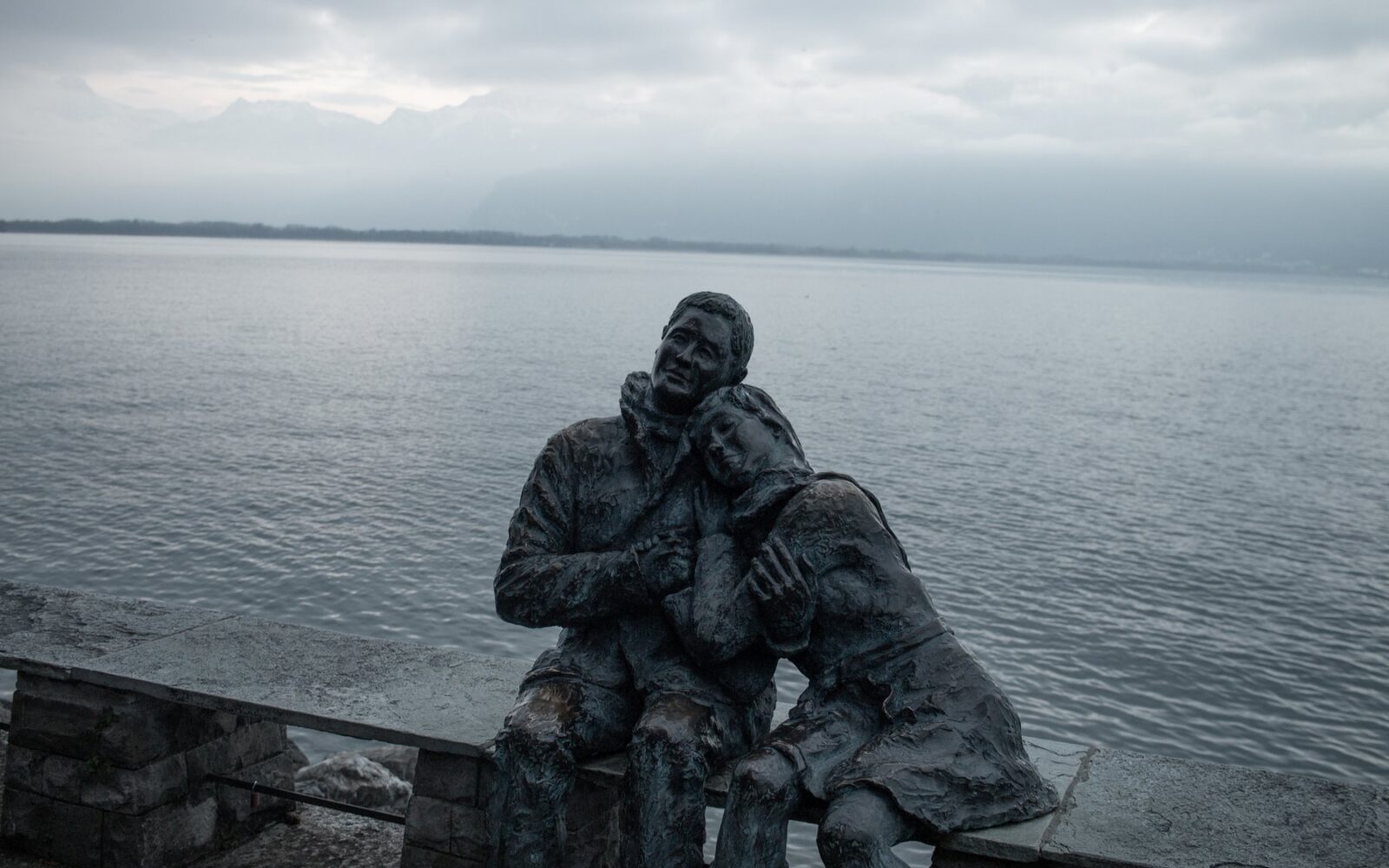 Escultura de dos figuras abrazadas frente a un mar gris al anochecer