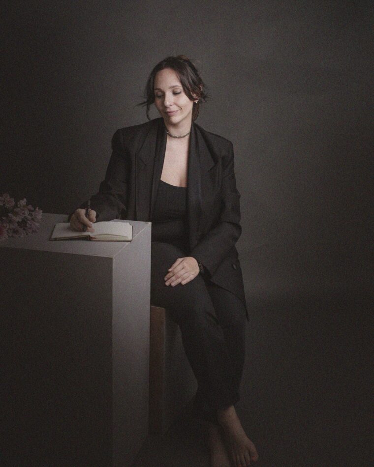Mía García, vestida de traje oscuro, escribiendo en un cuaderno apoyada en un pedestal